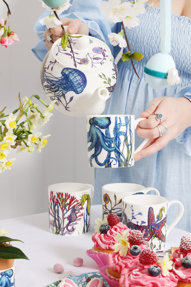 Table set for Easter with cupcakes and spring flowers. Through the blossom you can see a girl in a pale blue dress pouring tea into a cup, with both decorated in our Reef design of brightly coloured sea creatures, coral and seaweeds.