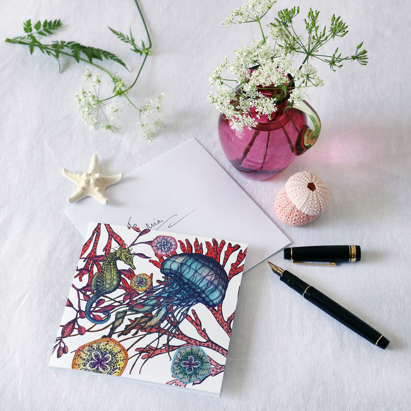 brightly coloured greeting card with jellyfish, seahorse and sea coral with baby jellyfish on a white background lying on a white table cloth with a fountain pen, hand written envelope shells and a small cranberry glass jug with wild flowers in 