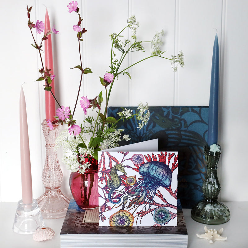 brightly coloured greeting card with jellyfish, seahorse and sea coral with baby jellyfish on a white background on shelf with pink and blue candles in candlesticks and a small cranberry glass jug with wild flowers in