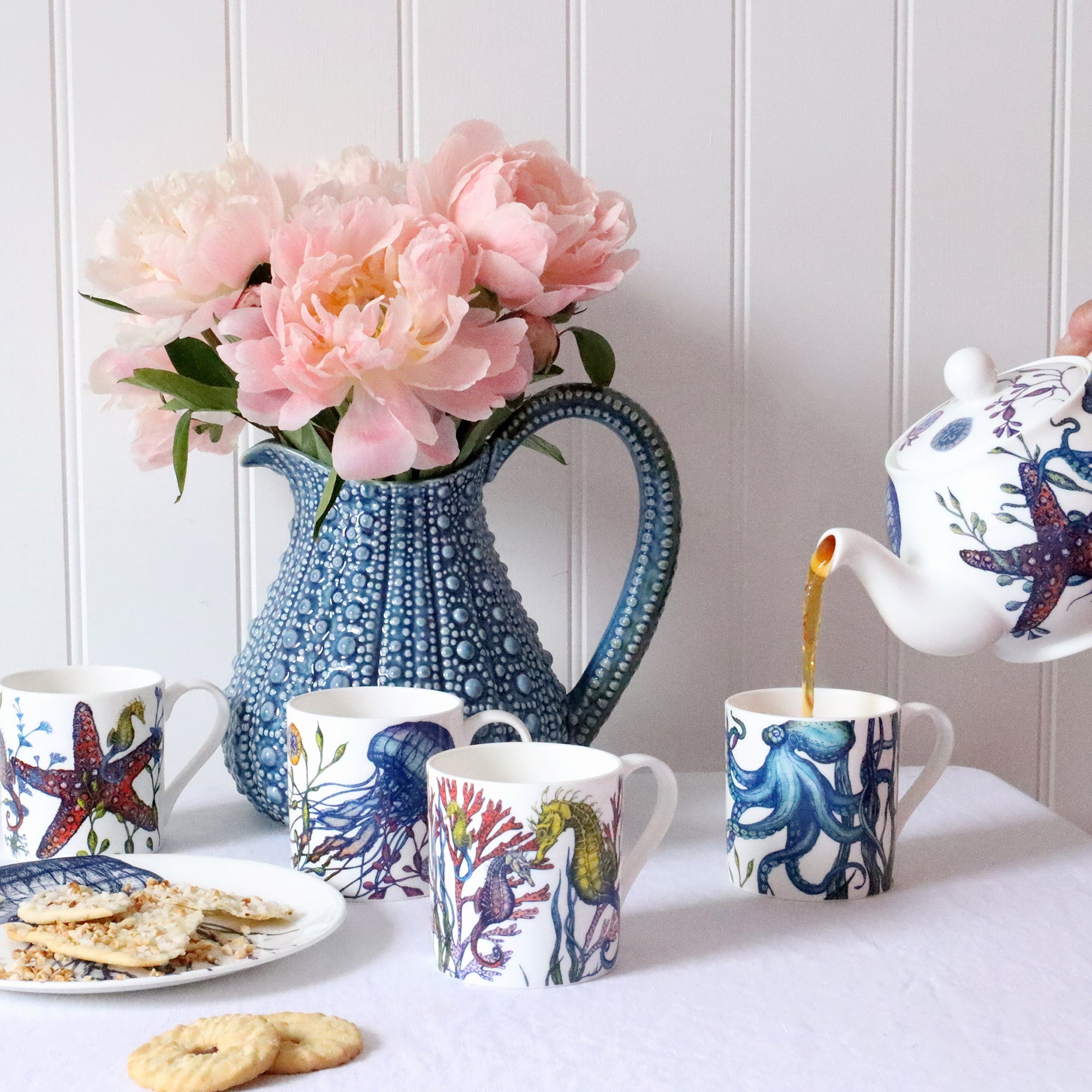 Set of four Bone china white mugs featuring hand drawn Reef designs stacked showing each design.One Starfish,Octopus,Seahorse and a Jellyfish in our bright reef colours with a reef teapot pouring tea into the reef octopus mug.