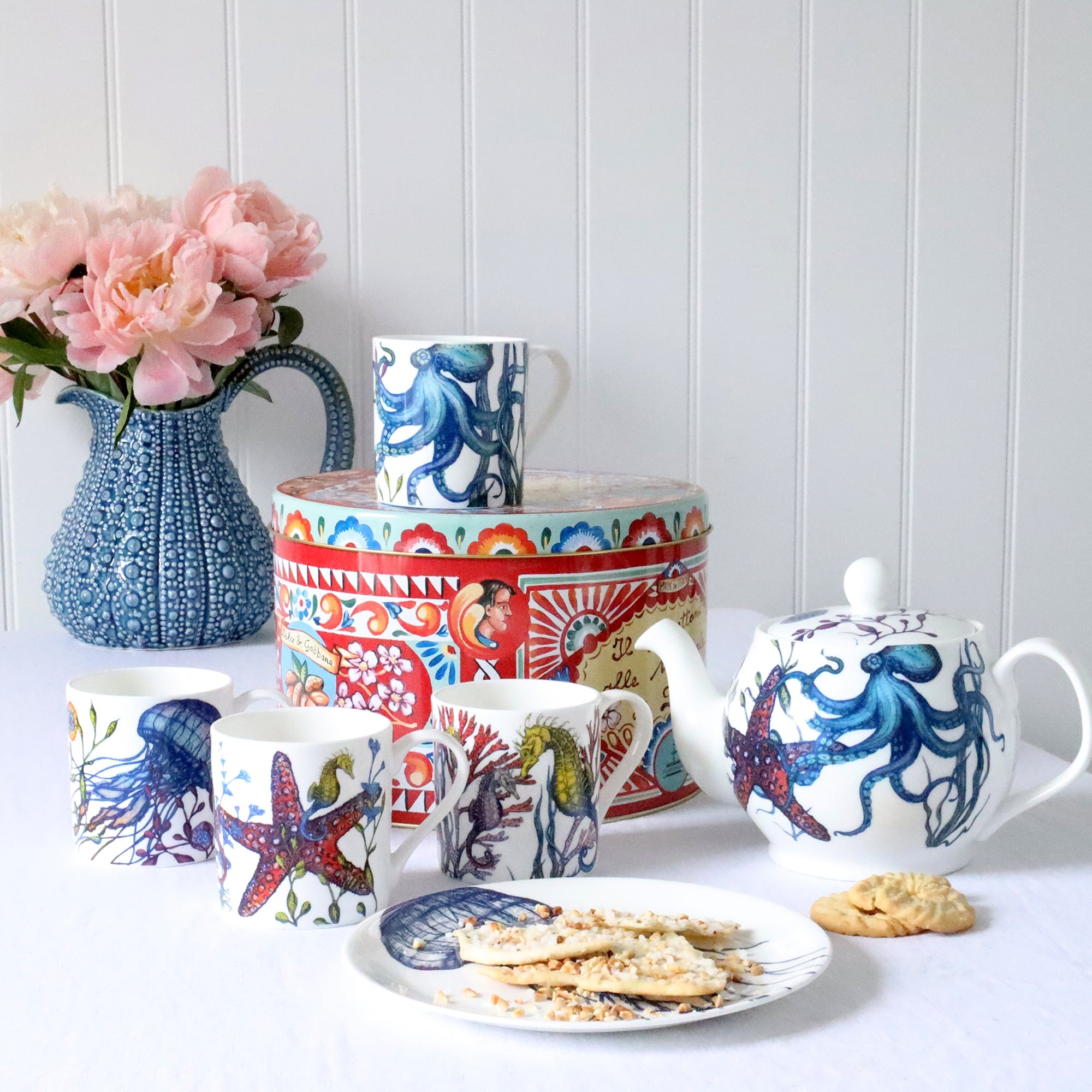 Set of four Bone china white mugs featuring hand drawn Reef designs stacked showing each design.One Starfish,Octopus,Seahorse and a Jellyfish in our bright reef colours on a white table setting with urchin jug, flowers and panettone tin.
