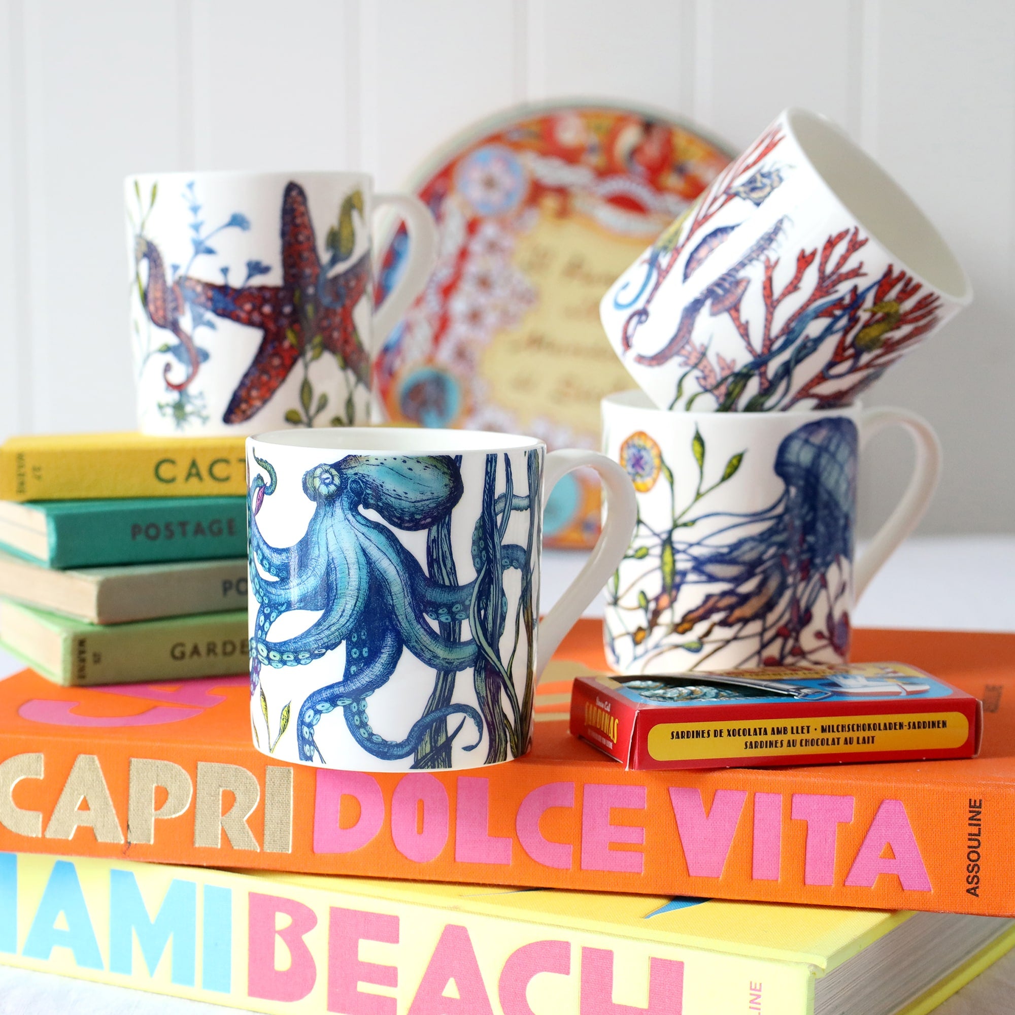 Set of four Bone china white mugs featuring hand drawn Reef designs stacked showing each design.One Starfish,Octopus,Seahorse and a Jellyfish in our bright reef colours all sitting on brightly coloured books.