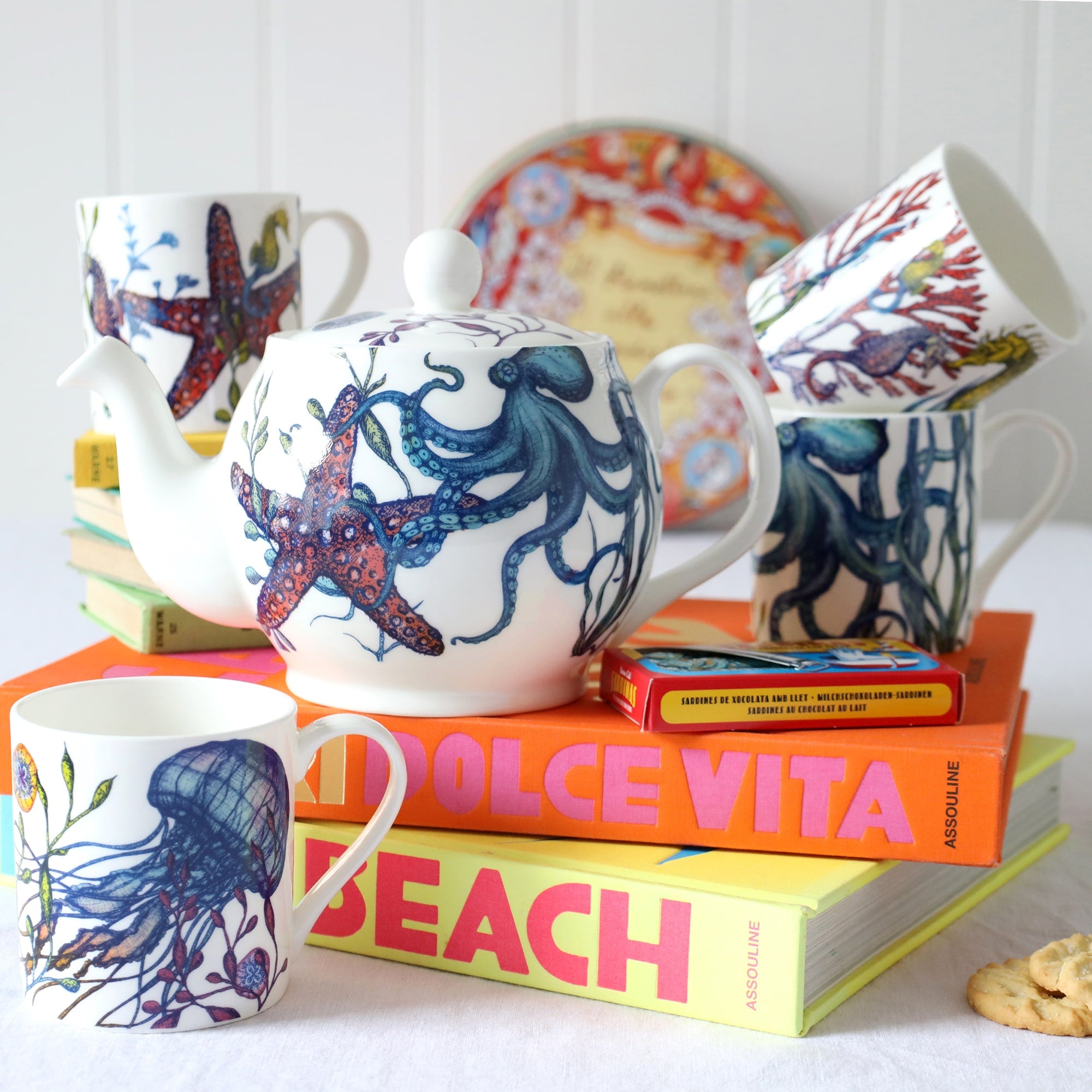 Set of four Bone china white mugs & teapot featuring hand drawn Reef designs stacked showing each design.One Starfish,Octopus,Seahorse and a Jellyfish in our bright reef colours all sitting on brightly coloured books.