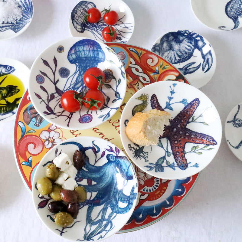 Nibbles bowl in Bone China in our colourful Reef range in the Jellyfish design placed on a brightly coloured tin with other nibbles bowls from our Reef range.All bowls have olives,bread ,tomatoes in.In the background you can see other items on the table