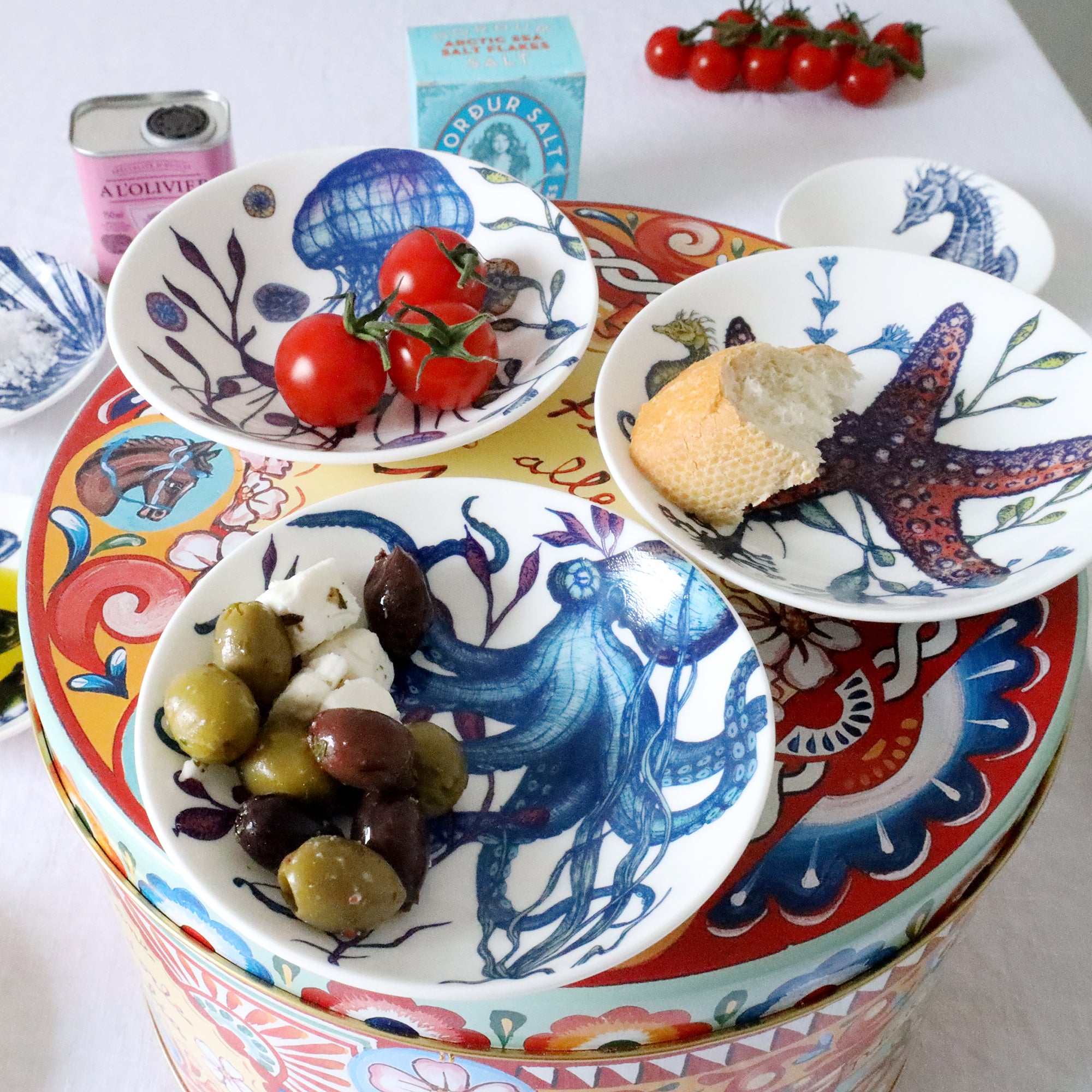 Nibbles bowl in Bone China in our colourful Reef range in the Jellyfish design placed on a brightly coloured tin with other nibbles bowls from our Reef range.All bowls have olives,bread ,tomatoes in.In the background you can see other items on the table