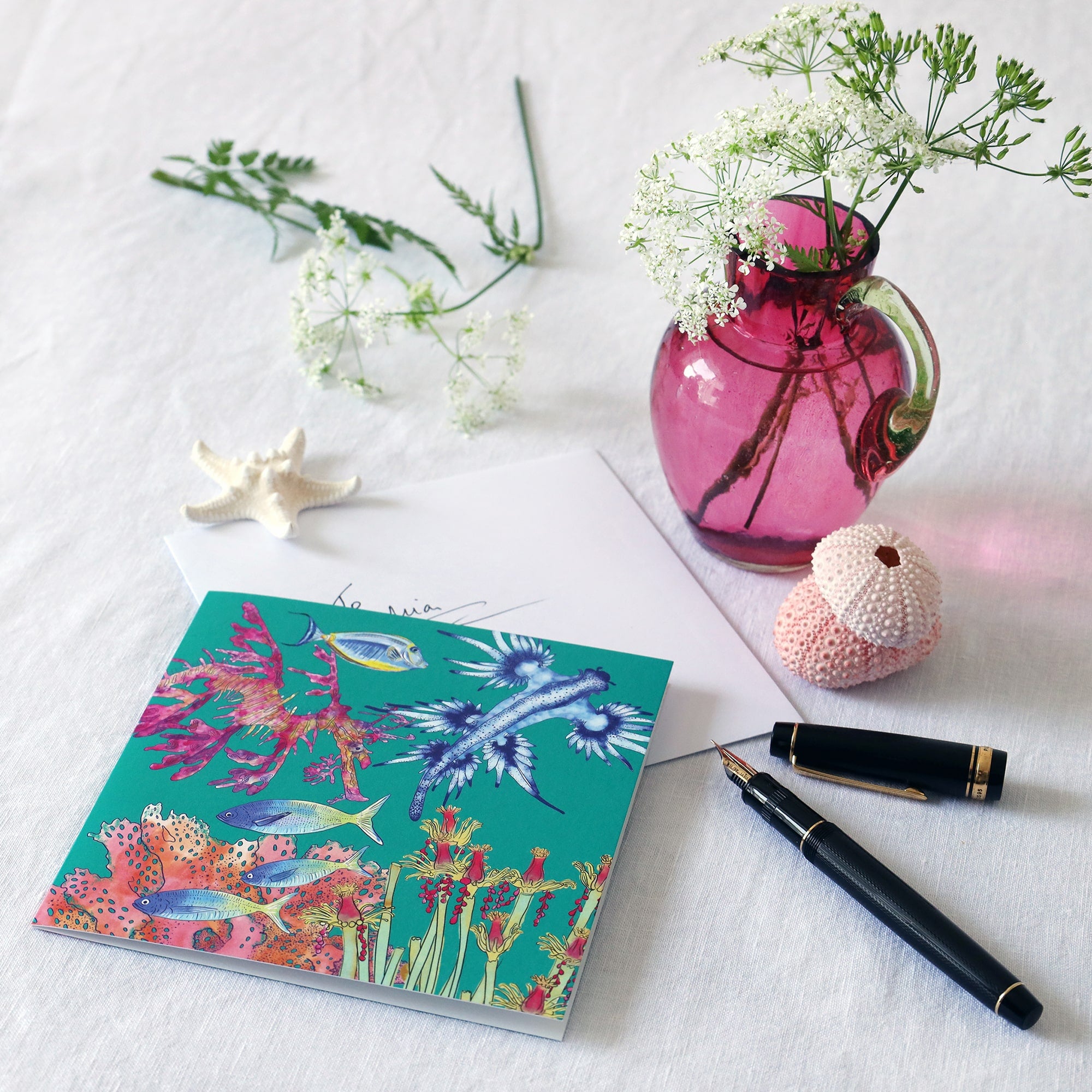 brightly coloured greeting card with illustrations of nudibranch, sea slug tropical fish and coral on a jade coloured background lying on a white table cloth with a fountain pen, hand written envelope shells and a small cranberry glass jug with wild flowers in