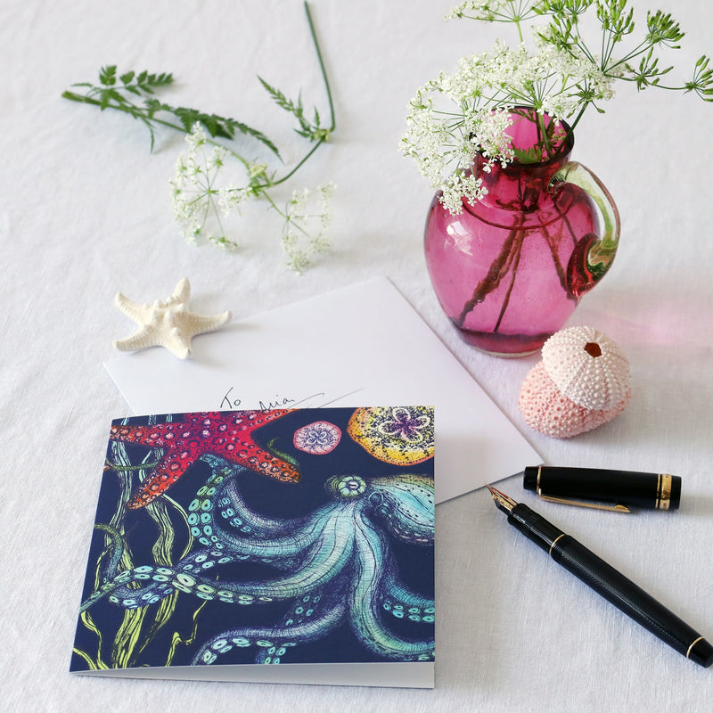 brightly coloured greeting card with octopus, starfish seaweed and baby jellyfish on a dark navy background lying on a white table cloth with a fountain pen, hand written envelope shells and a small cranberry glass jug with wild flowers in 