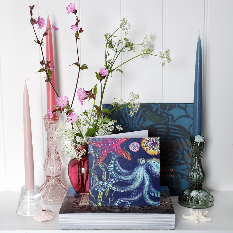 brightly coloured greeting card with octopus, starfish seaweed and baby jellyfish on a dark navy background on shelf with pink and blue candles in candlesticks and a small cranberry glass jug with wild flowers in 