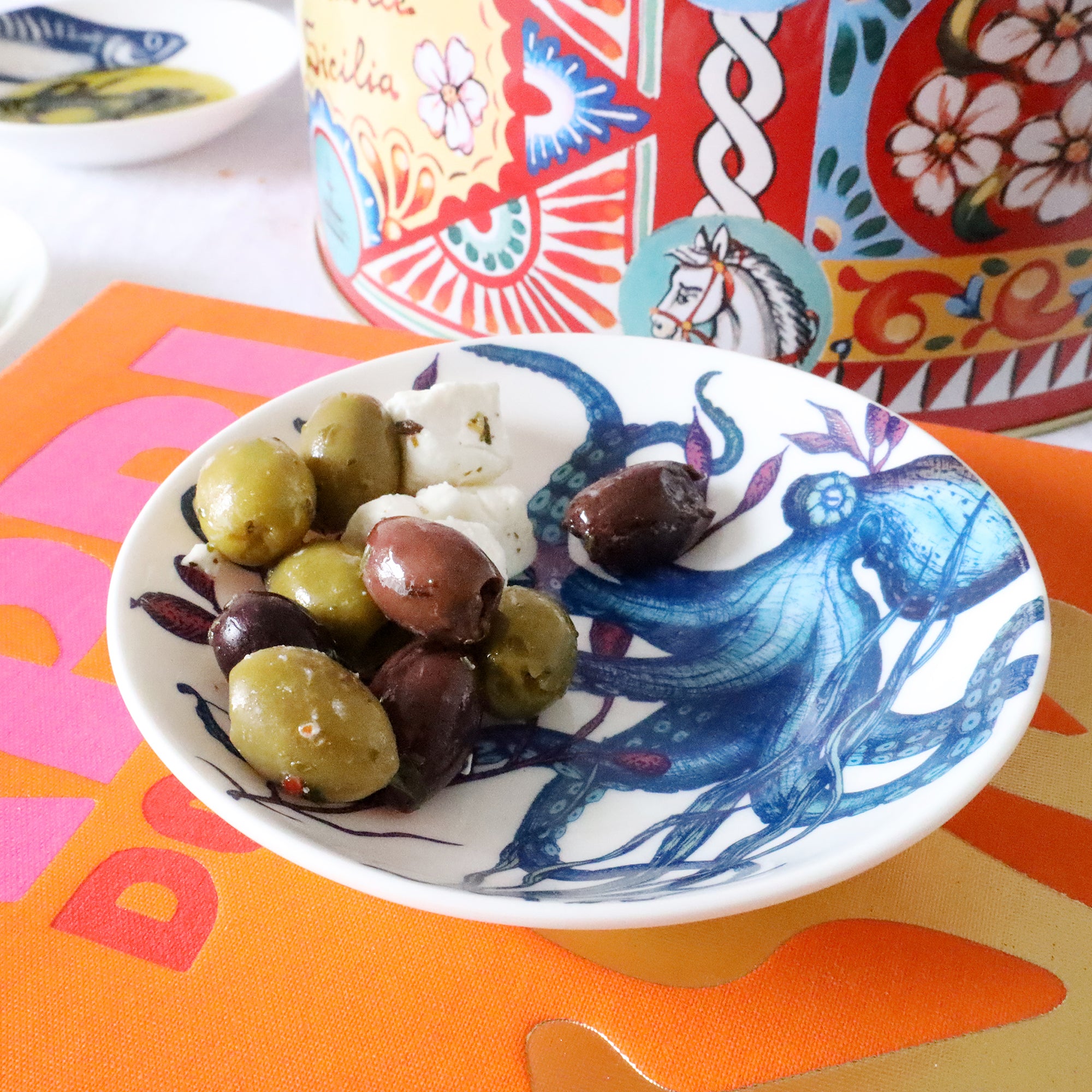 Nibbles bowl in Bone China in our colourful Reef range in the Octopus design with olives placed on a brightly coloured book.Next to it is a brightly coloured large tin and another reef nibbles bowl with oil in