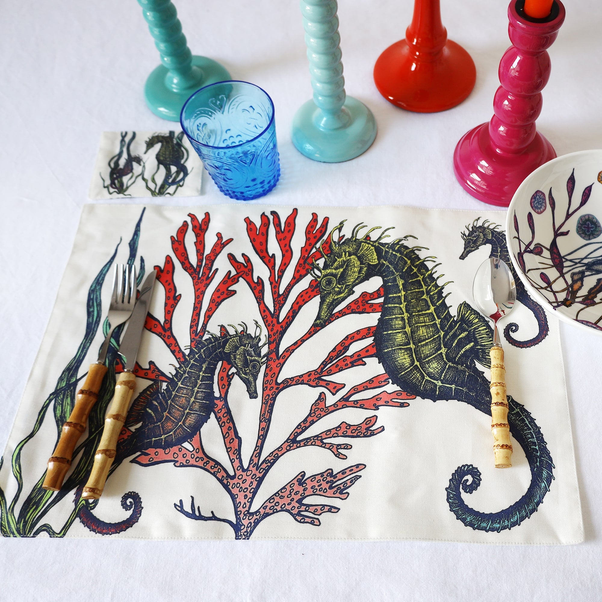 Fabric coaster in our Reef design showing two seahorses placed next to a matching placemat on a tablecloth.Also you can see bamboo cutlery.Behind are several coloured candle holders and a glass