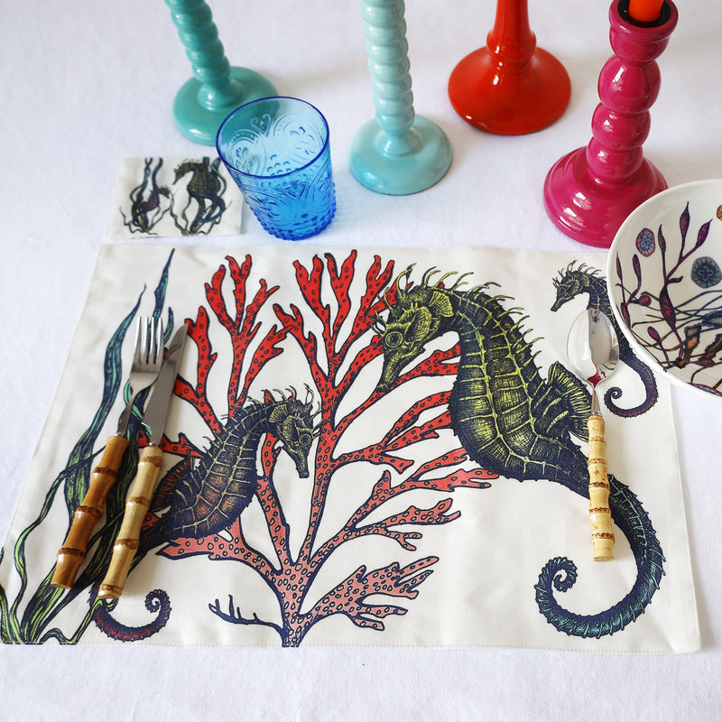 Cotton and linen mix white fabric rectangular placemats.Design is on both sides hand drawn illustrations set of four,showing the seahorse one on a white tablecloth. Adjacent is the matching coaster,on the placemat is our bamboo cutlery and a reef design bowl,in the background are several coloured candleholders