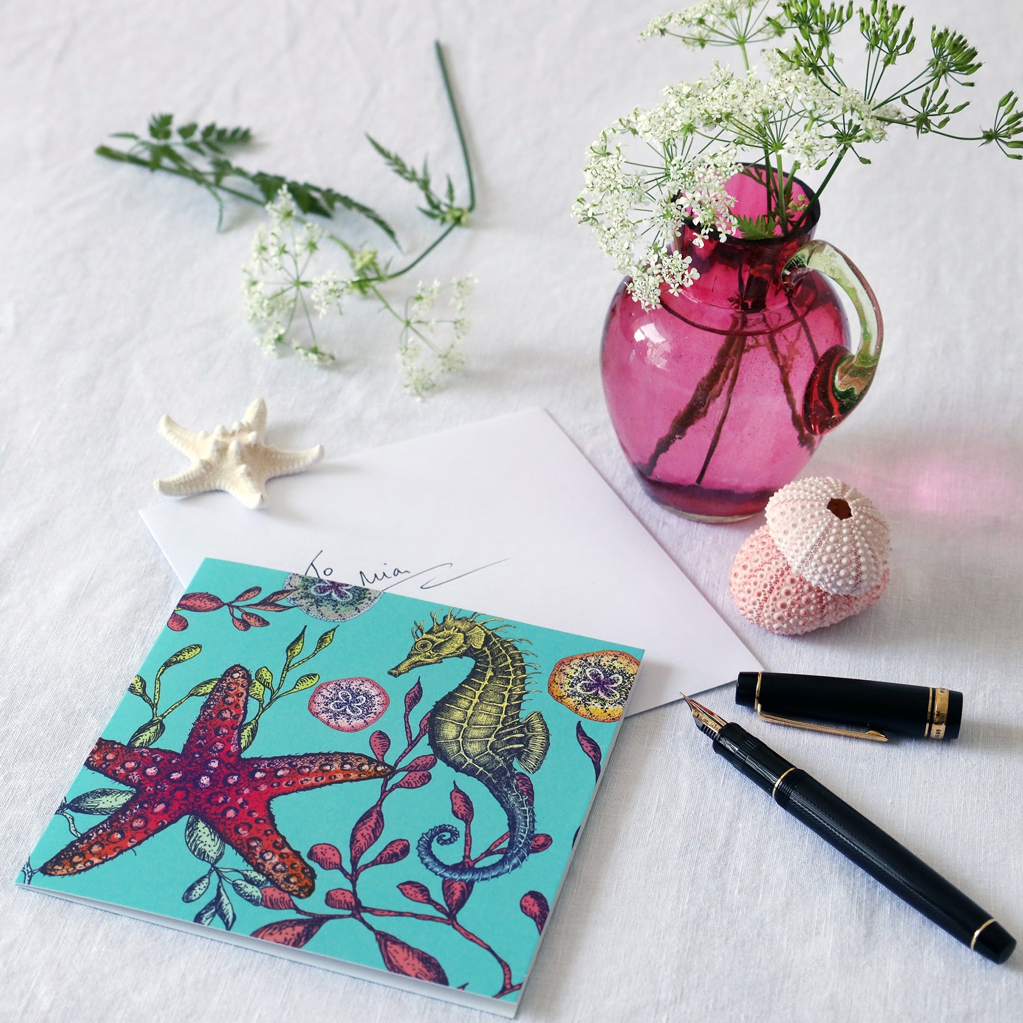 brightly coloured illustrated greeting card with seahorse, seaweeds, baby jellyfish and starfish on an aqua coloured background lying on a white table cloth with a fountain pen, hand written envelope shells and a small cranberry glass jug with wild flowers in 