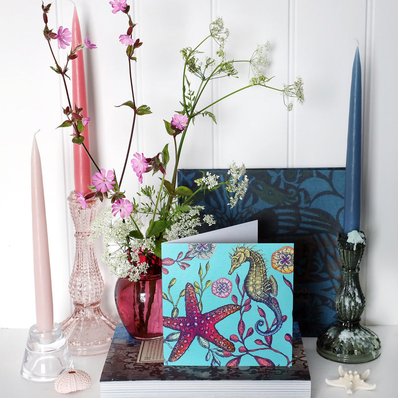 brightly coloured illustrated greeting card with seahorse, seaweeds, baby jellyfish and starfish on an aqua coloured background on shelf with pink and blue candles in candlesticks and a small cranberry glass jug with wild flowers in