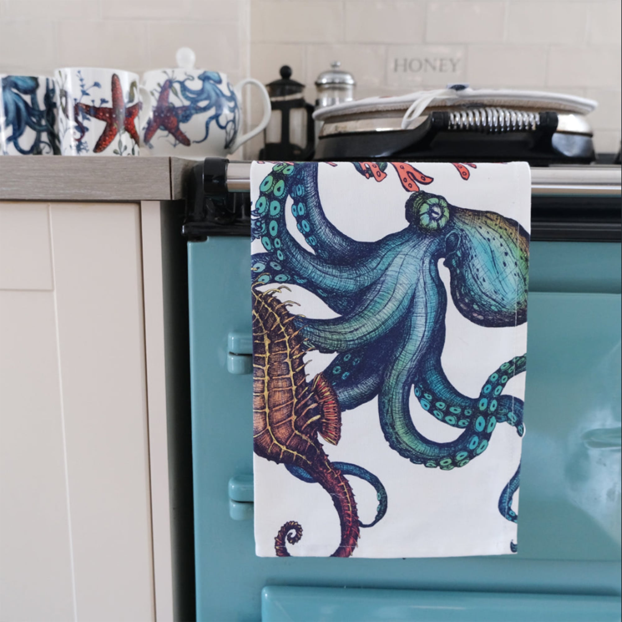 Cotton and Linen mix Tea Towel with hand drawn illustrations in Reef design draped over the arm of a green aga in a kitchen.On the kitchen worktop is a Reef teapot and matching mugs