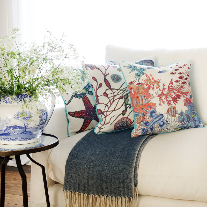 3 cushions an a white sofa with brightly coloured sea creatures illustrated  on with the sun streaming through the window at the back and a large willow pattern jug filled with cow parsley