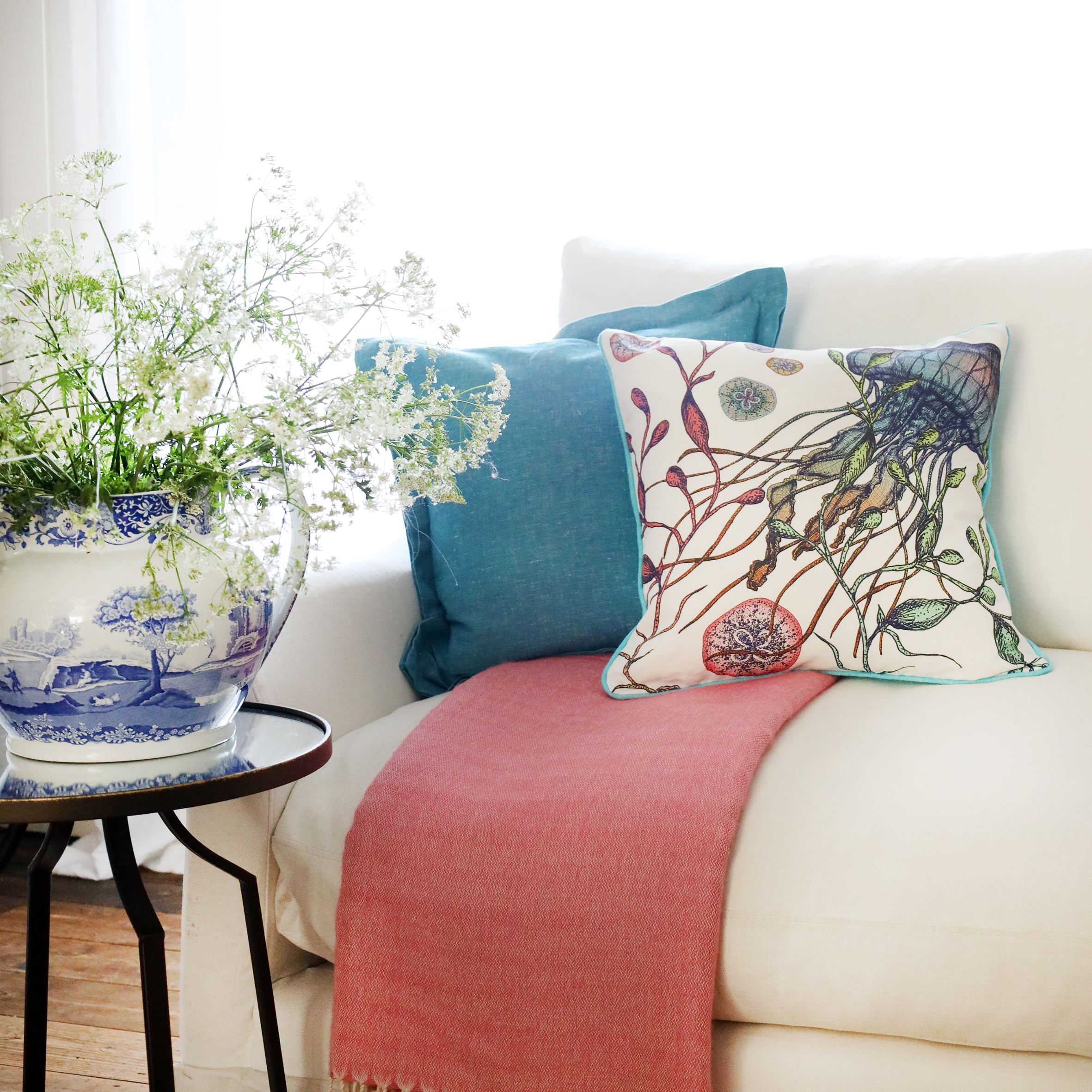 2 cushions an  a white sofa with brightly coloured jellyfish illustration on with the sun streaming through the window at the back and a large willow pattern jug filled with cow parsley