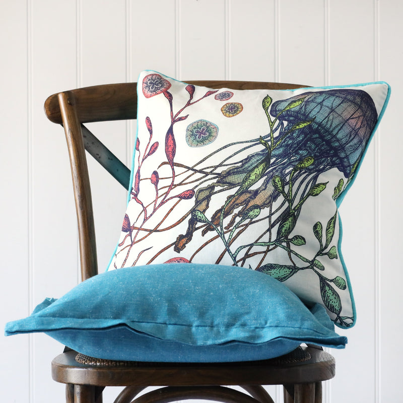white cushion with brightly coloured illustration of a jellyfish on the front, placed on a turquoise linen cushion on a wooden chair