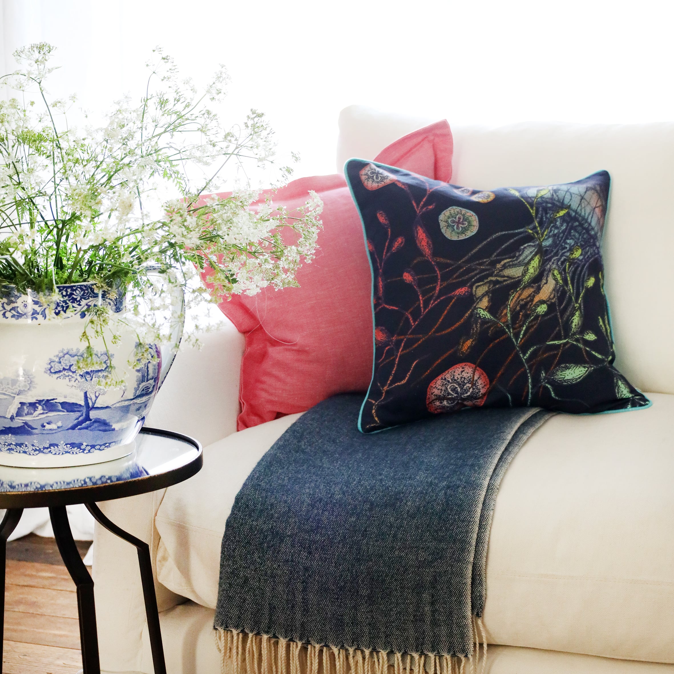 2 cushions an a white sofa with brightly coloured jellyfish illustration on a navy ground, with the sun streaming through the window at the back and a large willow pattern jug filled with cow parsley
