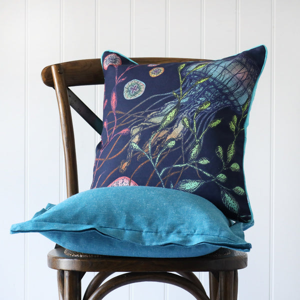 navy cushion with brightly coloured illustration of a jellyfish on the front, placed on a turquoise linen cushion on a wooden chair