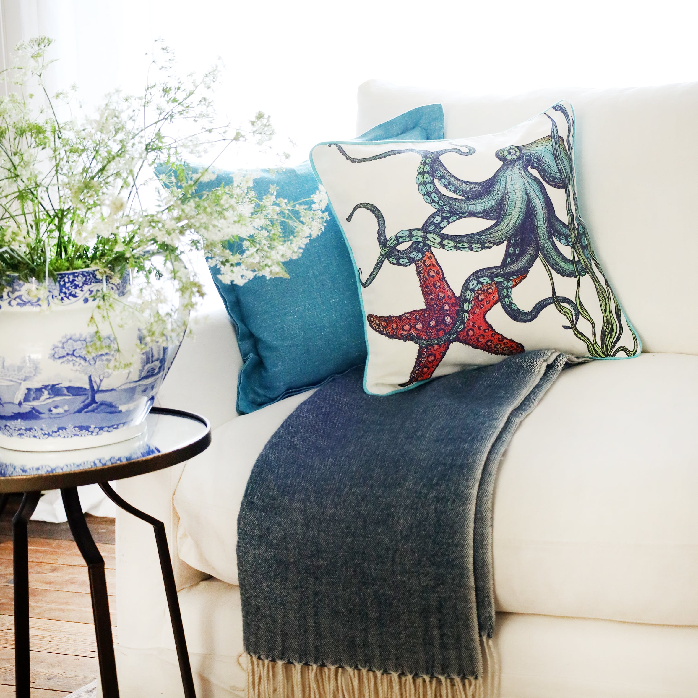 2 cushions an a white sofa with brightly coloured sea creatures illustrated  on with the sun streaming through the window at the back and a large willow pattern jug filled with cow parsley