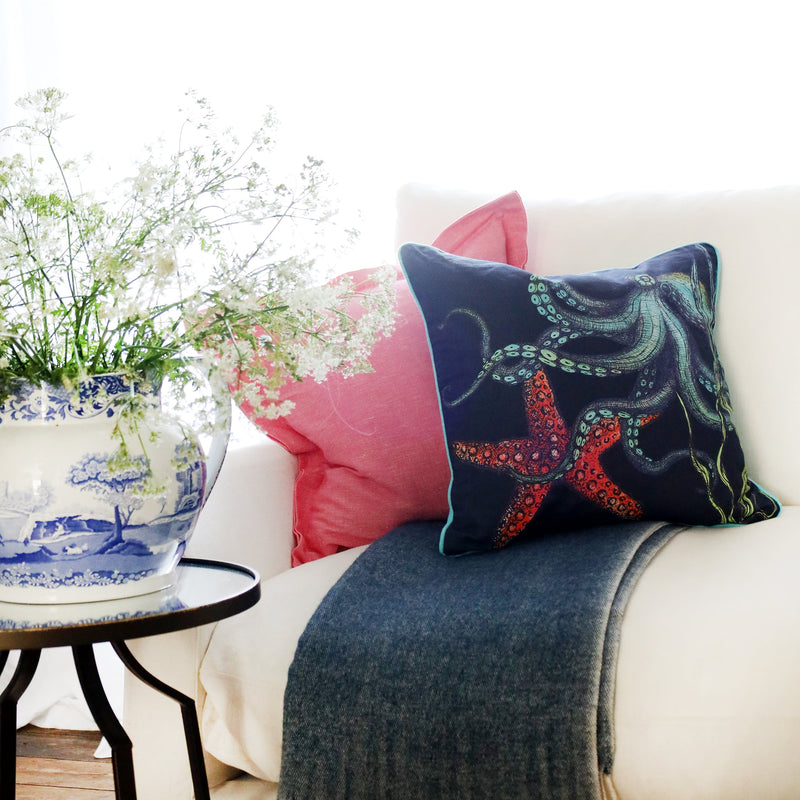 2 cushions an a white sofa with brightly coloured octopus and starfish illustration on a navy ground with the sun streaming through the window at the back and a large willow pattern jug filled with cow parsley
