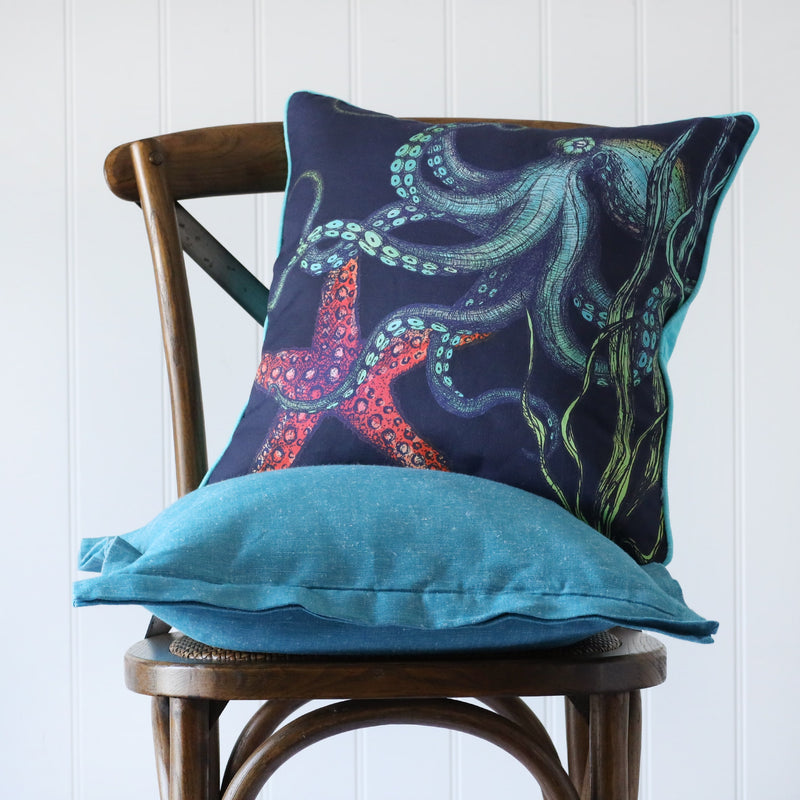 navy cushion with brightly coloured illustrated of an octopus and starfish on the front, placed on a turquoise linen cushion on a wooden chair
