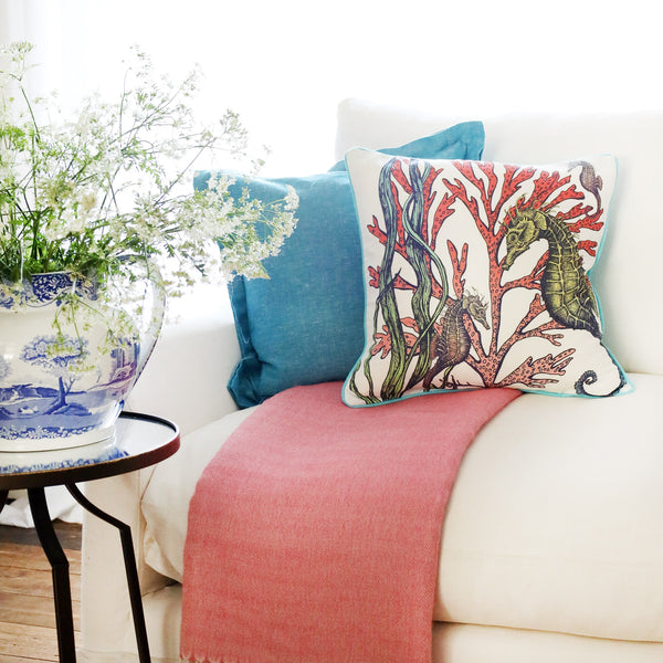 2 cushions an  a white sofa with brightly coloured seahorses and seaweed illustration on with the sun streaming through the window at the back and a large willow pattern jug filled with cow parsley