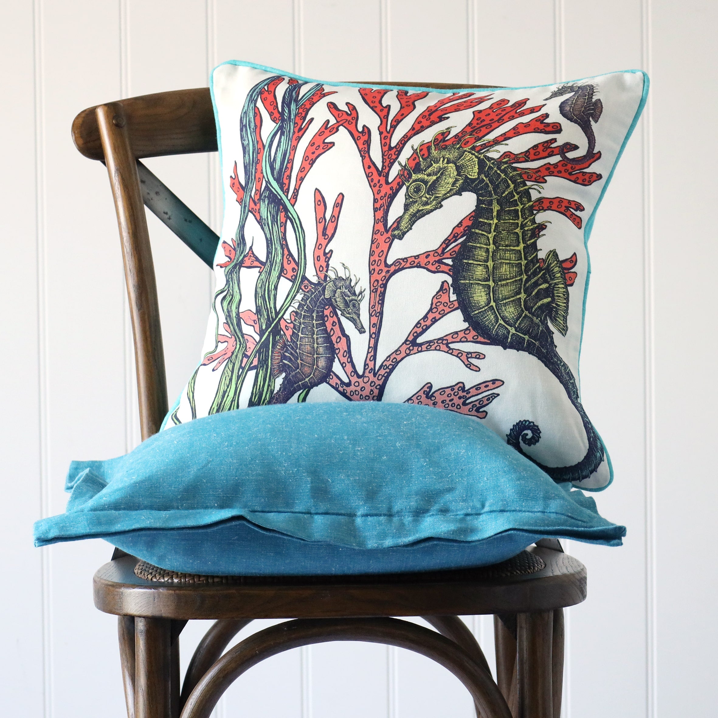 white cushion with brightly coloured illustration of seahorses and seaweeds on the front, placed on a turquoise linen cushion on a wooden chair