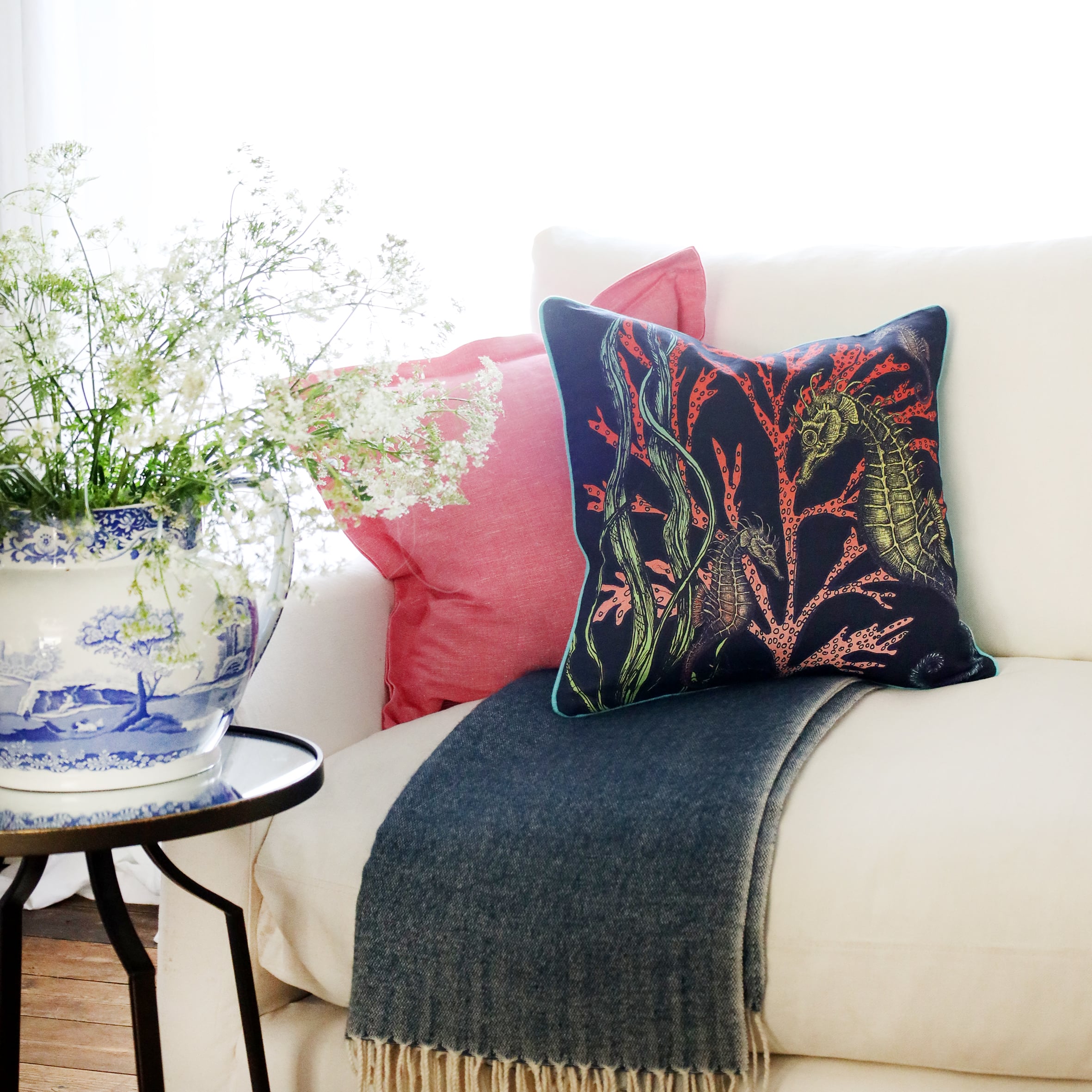2 cushions an  a white sofa with brightly coloured seahorses and seaweed illustration on a navy ground with the sun streaming through the window at the back and a large willow pattern jug filled with cow parsley