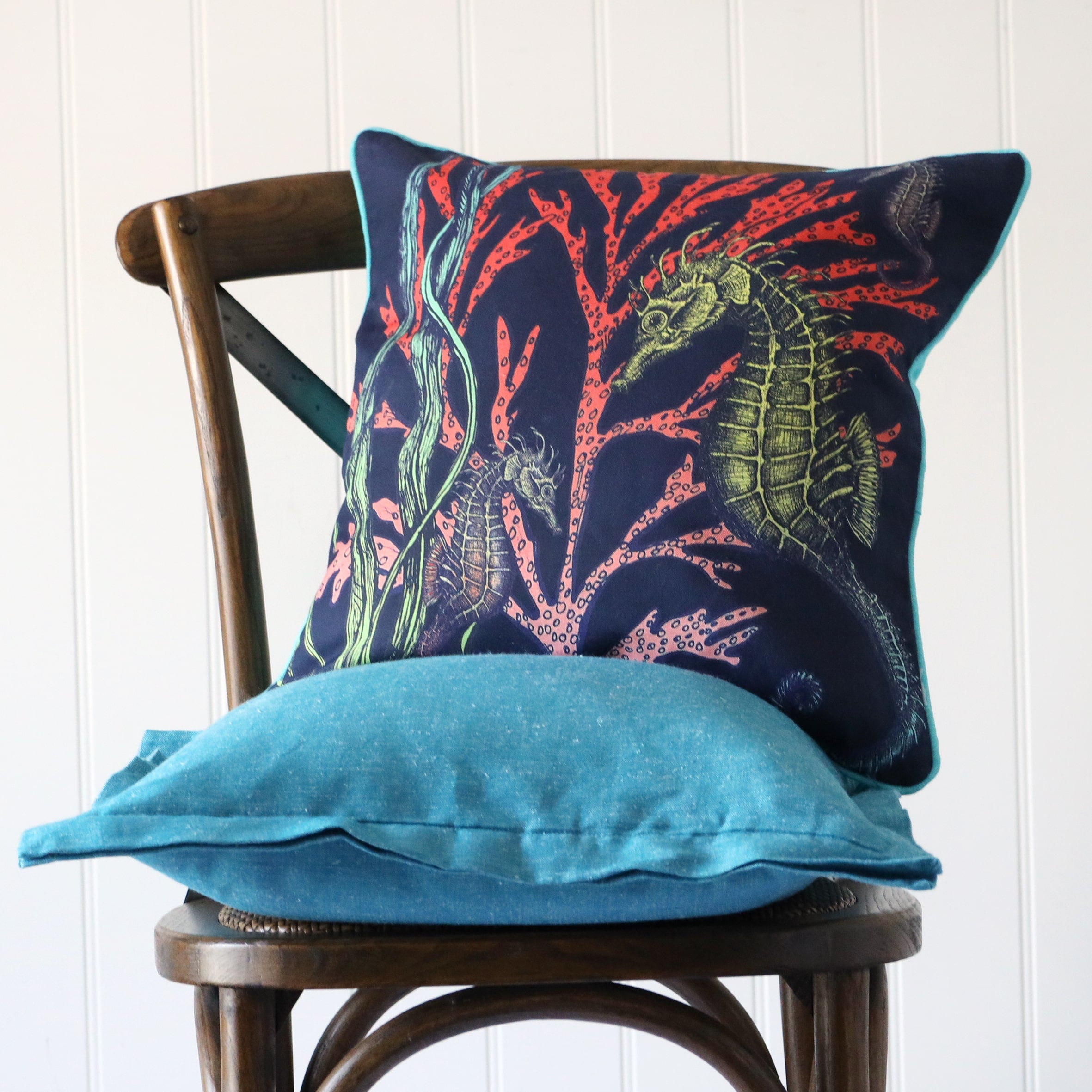 navy cushion with brightly coloured illustration of a seahorse and seaweed on the front, placed on a turquoise linen cushion on a wooden chair