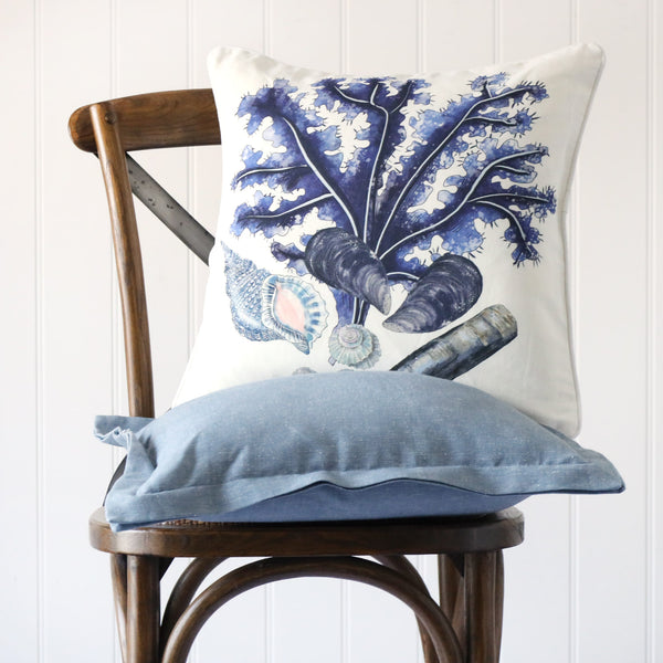 white cushion with illustrated shells and seaweed all in blue tones illustrated on the front, placed on a linen cushion on a wooden chair