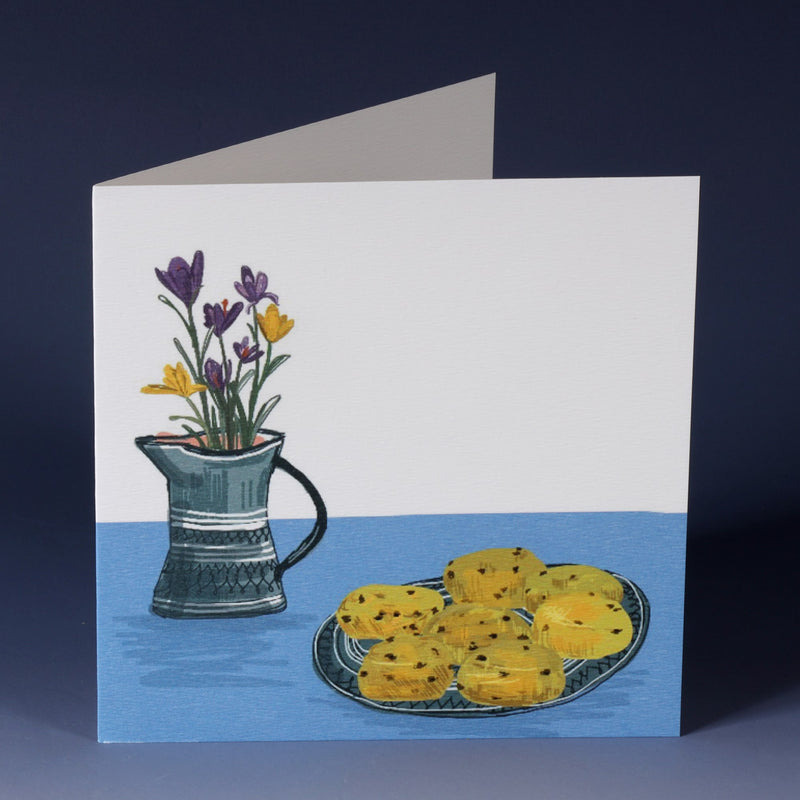 greeting card with an illustration of saffron buns  on a plate with a jug of crocus all sitting on a blue table cloth