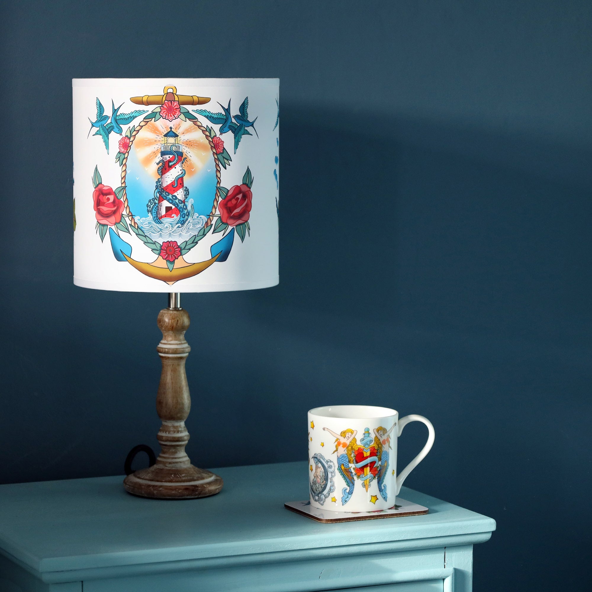 Small pixie whitewashed wooden lamp base with tattoo inspired lampshade with lighthouse and kraken design, with a mug with mermaids, heart and daggers design on the blue cabinet as well. All this is set against a dark denim blue wall.