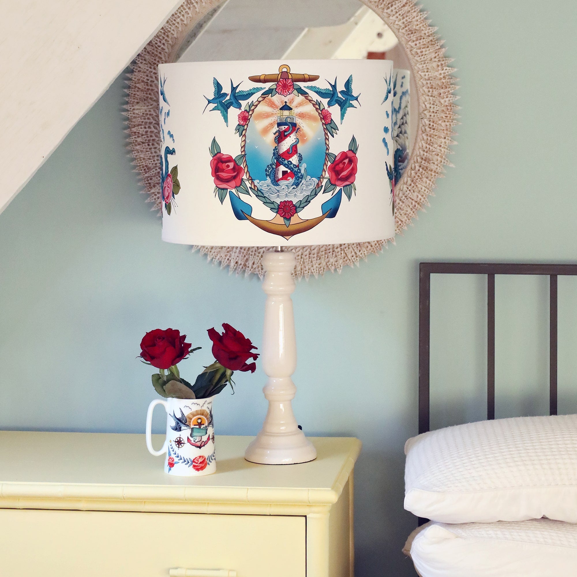 White bone china jug with brightly coloured tattoo inspired design of swallows, anchor and roses with red roses in it. This is sitting on a soft yellow set of drawers next to a bed. There is also a white lamp base with a tattoo inspired lampshade with a lighthouse, kraken, rope, anchor and roses.