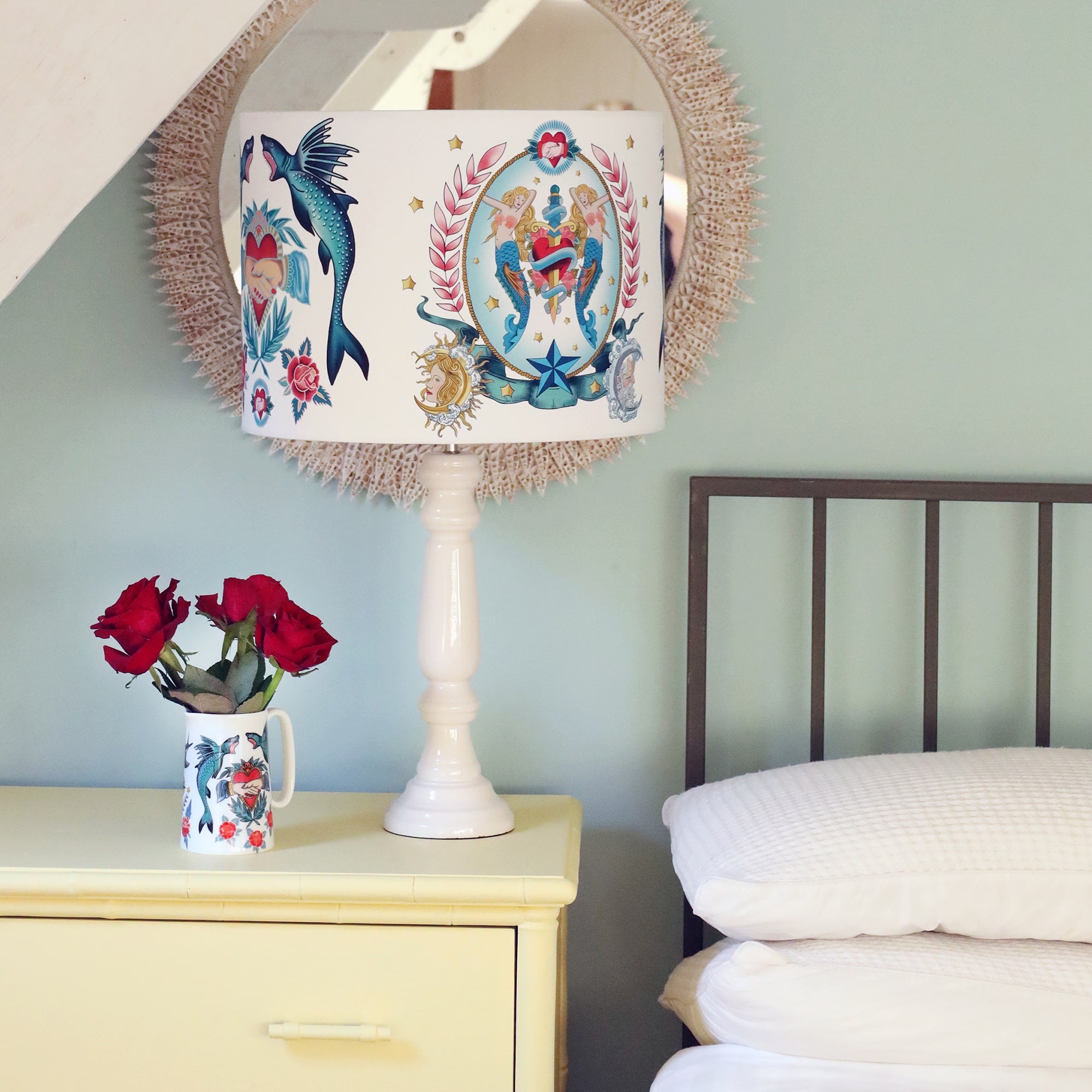 Soft yellow chest of drawers next to a bed with a glossy white lamp base and mermaids & daggers and sharks & heartstattoo inspired design. There is a small jug with swallows and anchor design with red roses next to the lamp.