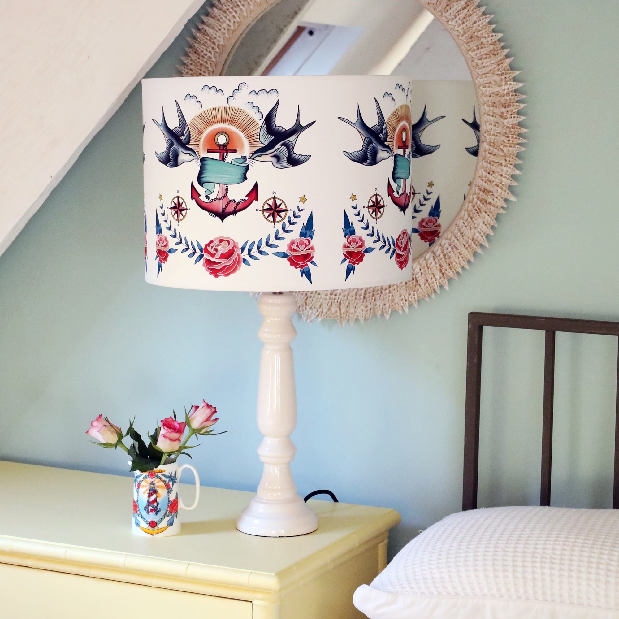 Soft yellow chest of drawers next to a bed with a glossy white lamp base and swallows, anchor and roses tattoo inspired design. There is a small jug with swallows and anchor design with red roses next to the lamp.