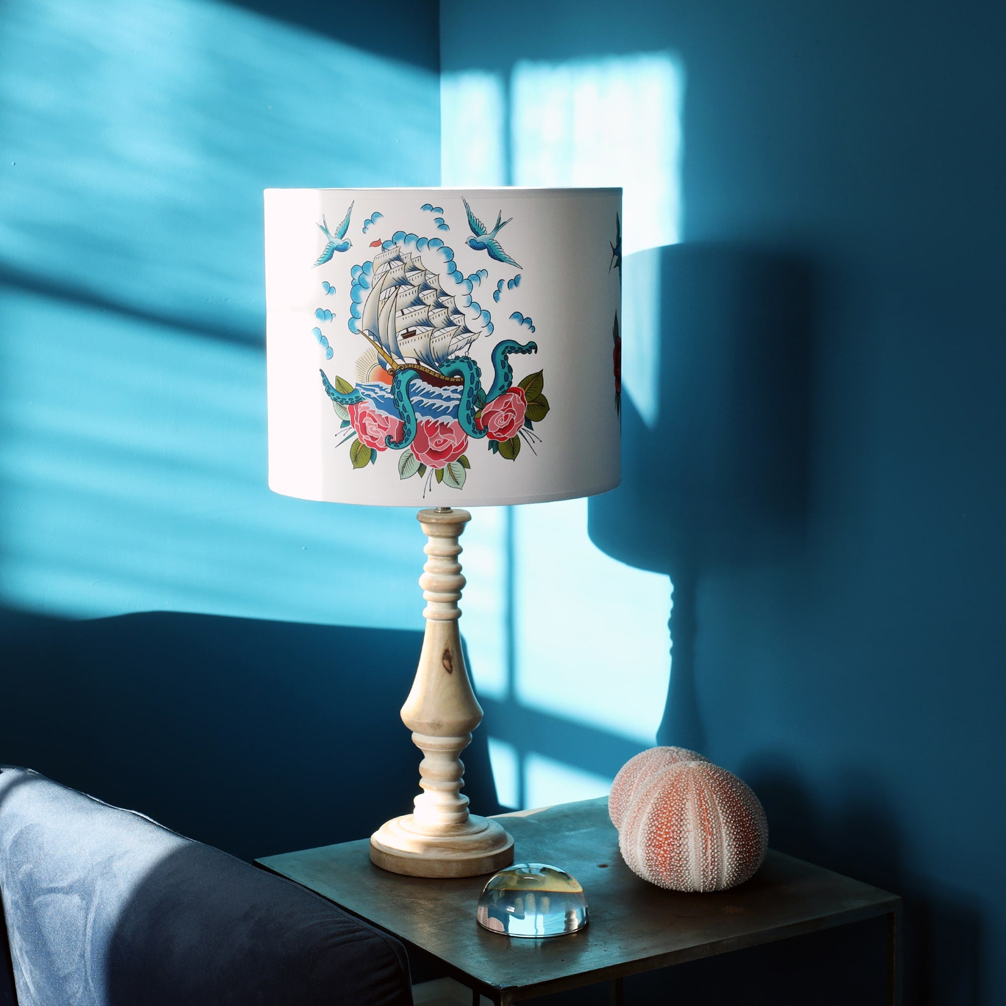 Table lamp in a teal sunny corner, the whitewashed wooden base is topped with a lampshade with ship, kraken and roses from our Sailor's story collection.