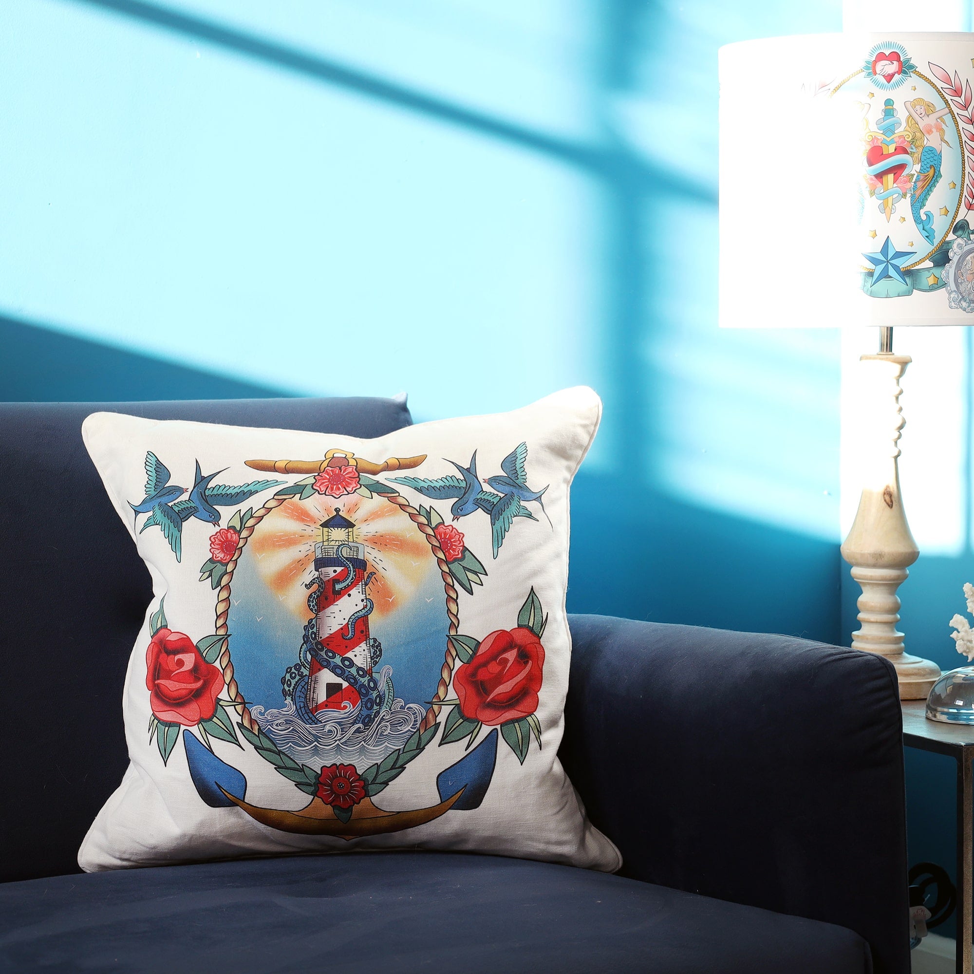 White cushion with brightly coloured tattoo inspired lighthouse and kraken design in a rope and anchor vignette with roses and swallows, on a navy velvet settee with sun streaming in.