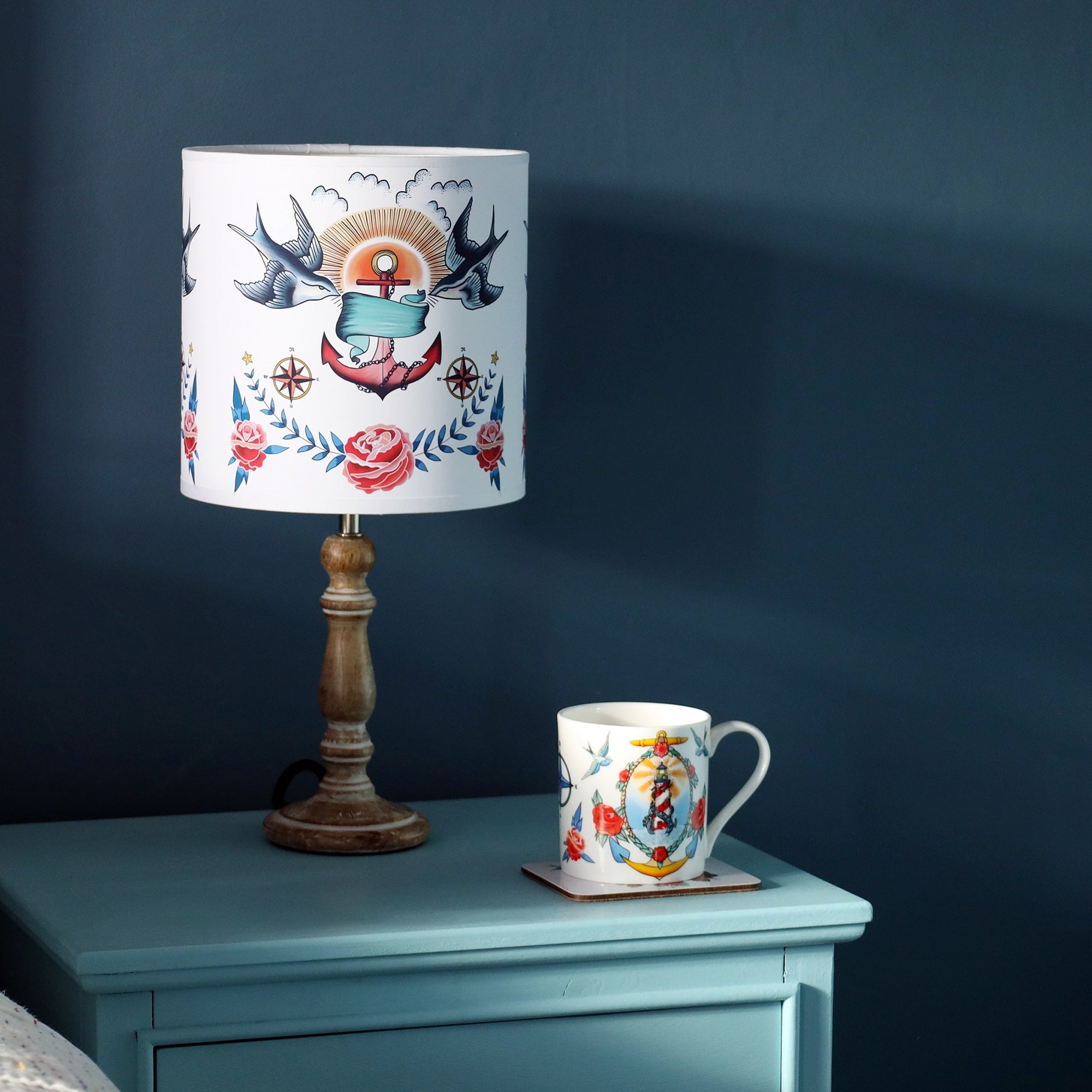 Small pixie whitewashed wooden lamp base with tattoo inspired lampshade with swallows, anchor and roses design, with a mug with lighthouse and kraken design on the blue cabinet as well. All this is set against a dark denim blue wall.