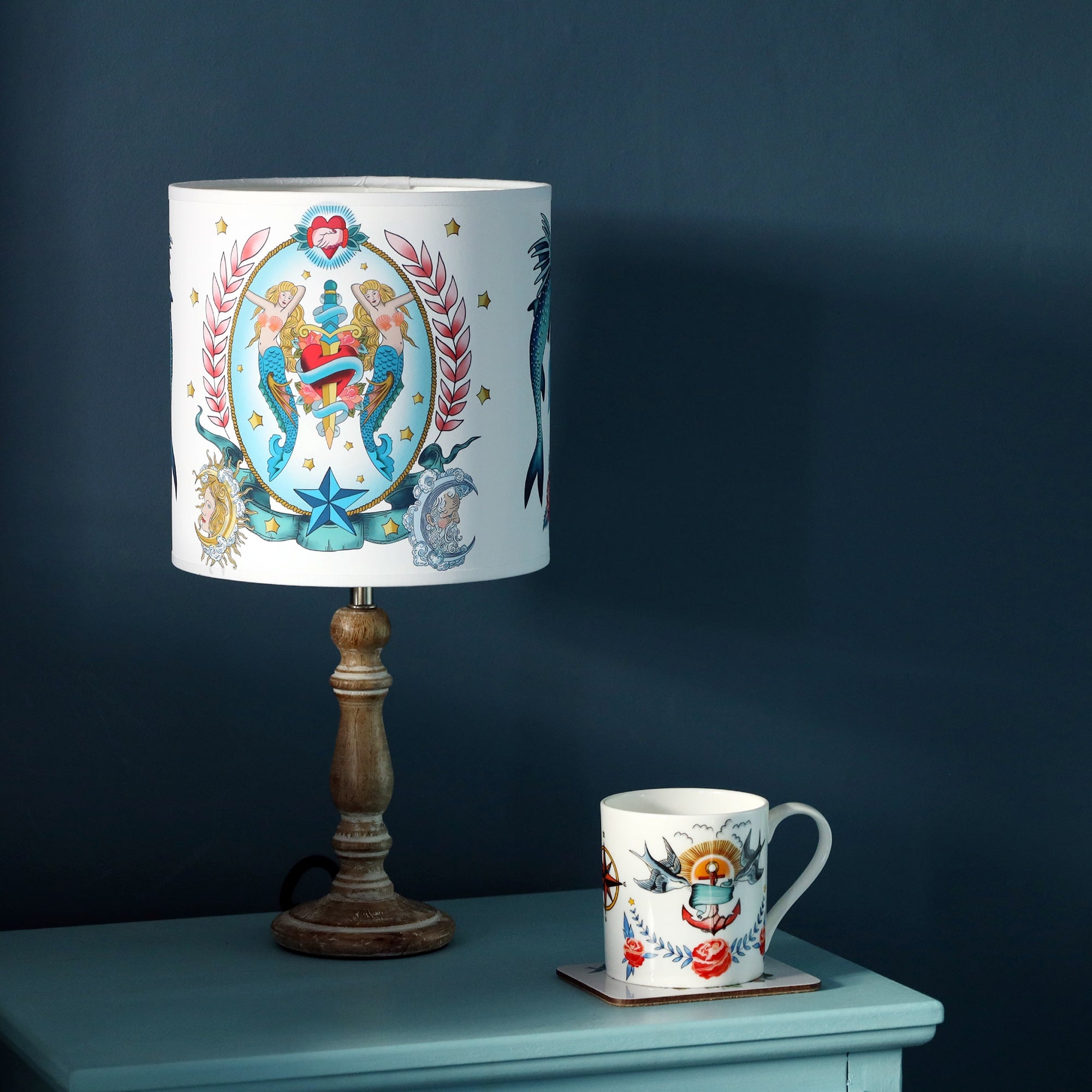 Small pixie whitewashed wooden lamp base with tattoo inspired lampshade with mermaids, heart and dagger design, with a mug with lighthouse and kraken design on the blue cabinet as well. All this is set against a dark denim blue wall.