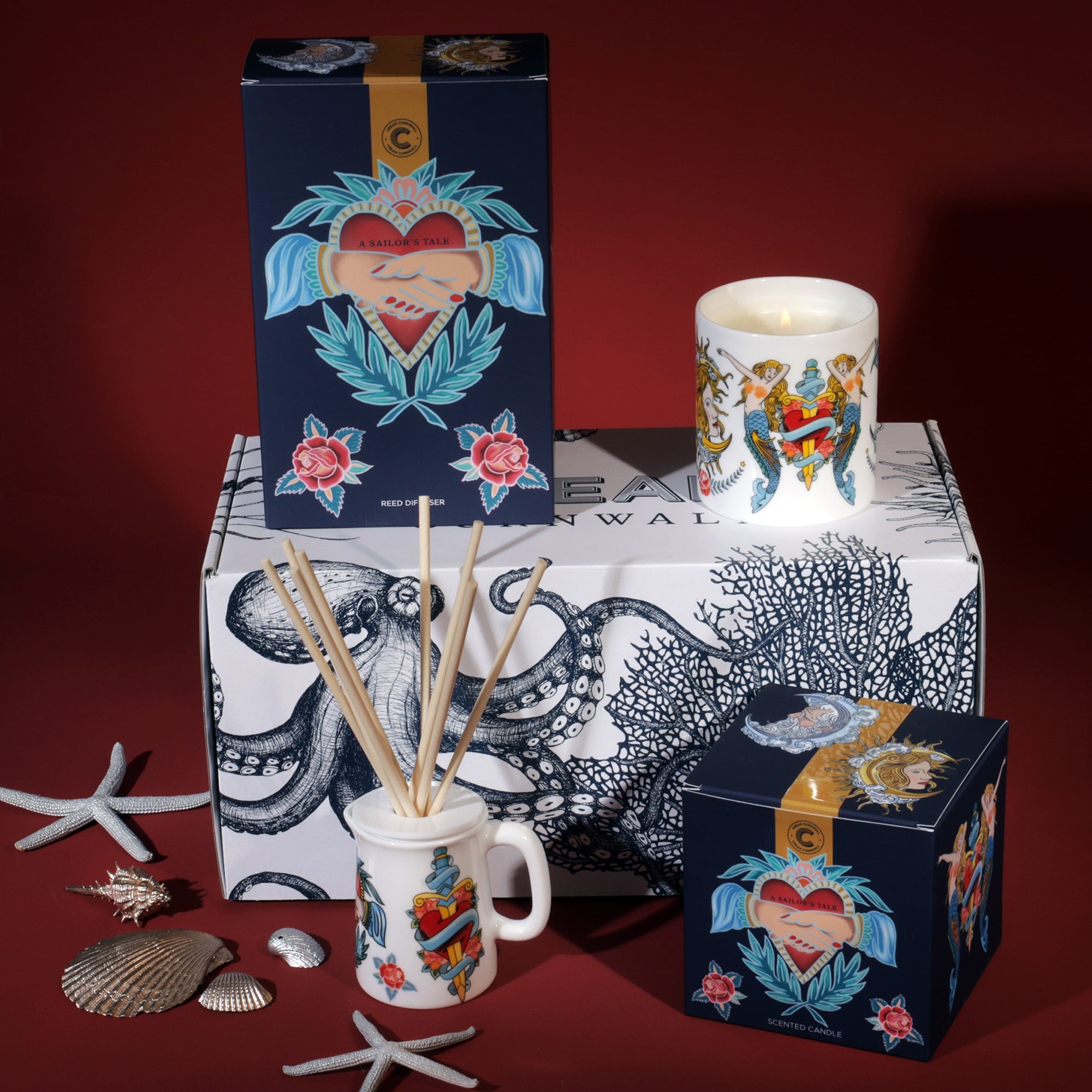 A display of Cream Cornwall fragrance products featuring a nautical theme. The setup includes a reed diffuser, a scented candle, and a gift box, all decorated with tattoo-style designs of hearts, roses, and anchors. The packaging is predominantly navy blue with vibrant red and teal accents. The background is a deep red, with decorative seashells and starfish placed around the products, enhancing the maritime aesthetic.