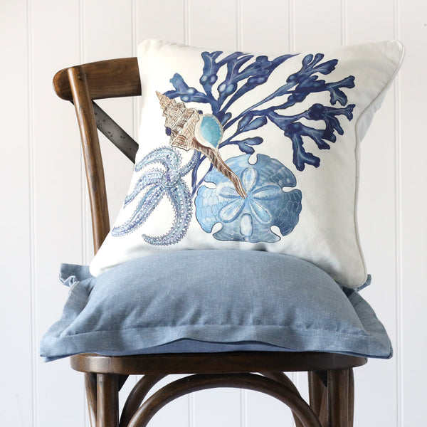 white cushion with illustrated shell, starfish, sand dollar and seaweed all in blue tones illustrated on the front, placed on a linen cushion on a wooden chair