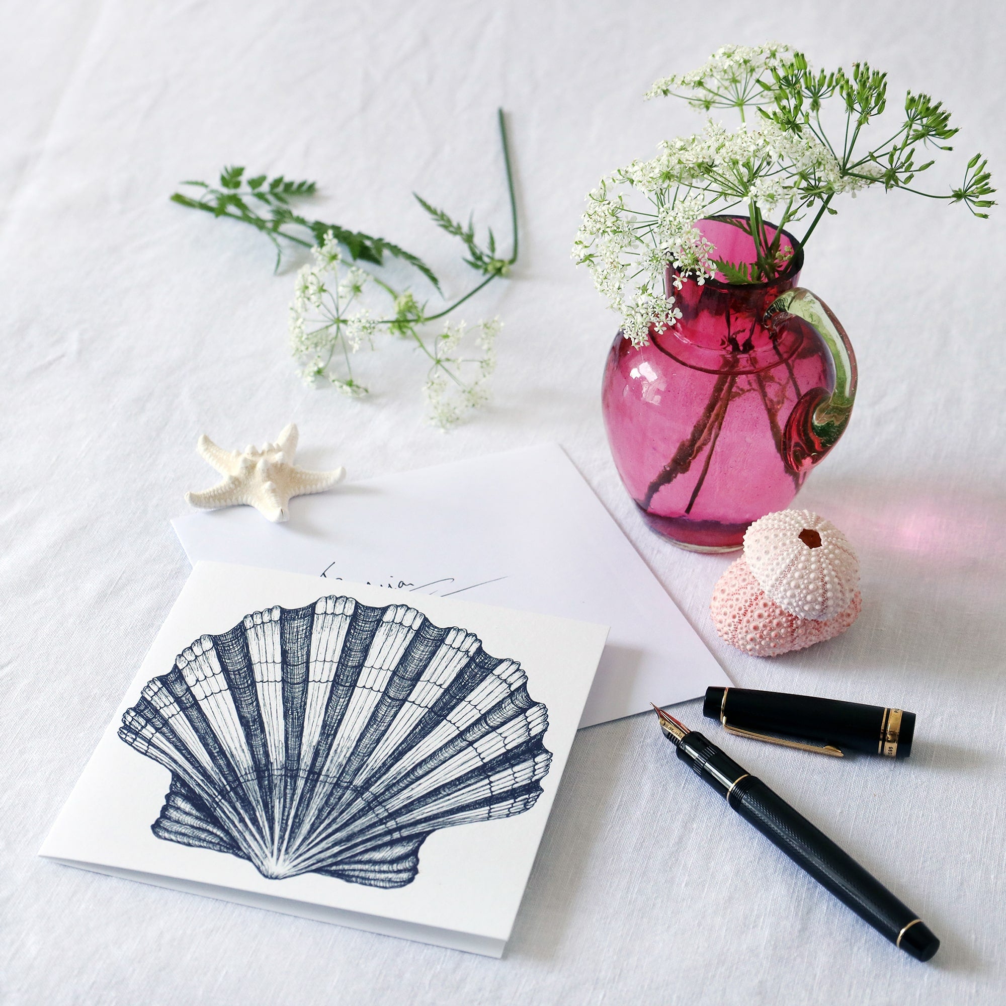 greeting card with navy illustration of a scallop shell on a white background lying on a white table cloth with a fountain pen, hand written envelope shells and a small cranberry glass jug with wild flowers in 