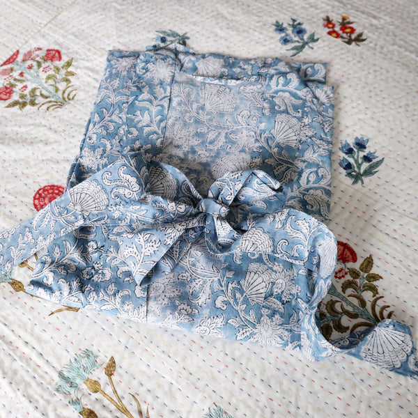 Seashell Flower dressing gown which is Hand block printed fabric in a soft blue and the print is swirling shells and tendrils in white.Placed on a bed with a white cover with occasional  bright flowers.