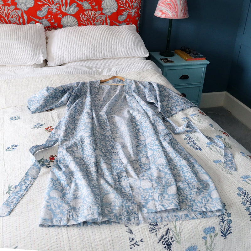 Seashell Flower dressing gown which is Hand block printed fabric .The gown is placed on a bed with a white cover with occasional bright flowers. You can see the blue bedside cabinet with a lamp base and a marble swirl lampshade.