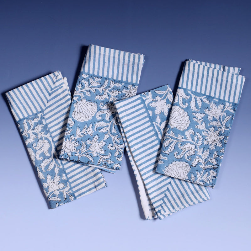 Seashell Flower napkins which are Hand block printed fabric in a soft blue and the print is swirling shells and tendrils in white on a napkin.Around the pattern is a white and blue striped finished edge.There are four of them on top of each other in a line.