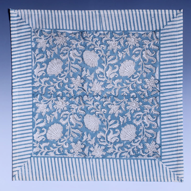 Seashell Flower napkins which are Hand block printed fabric in a soft blue and the print is swirling shells and tendrils in white on a napkin.Around the pattern is a white and blue striped finished edge
