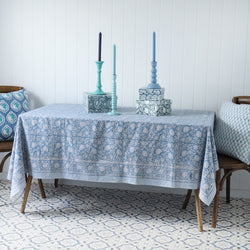 Seashell Flower tablecloth which is a Hand block printed fabric  .Around the pattern is a white and blue striped finished edge.It is placed on a rectangle table decorated with Hand decorated boxes and candlesticks.Either side of the table are chairs with a hand block printed cushion.