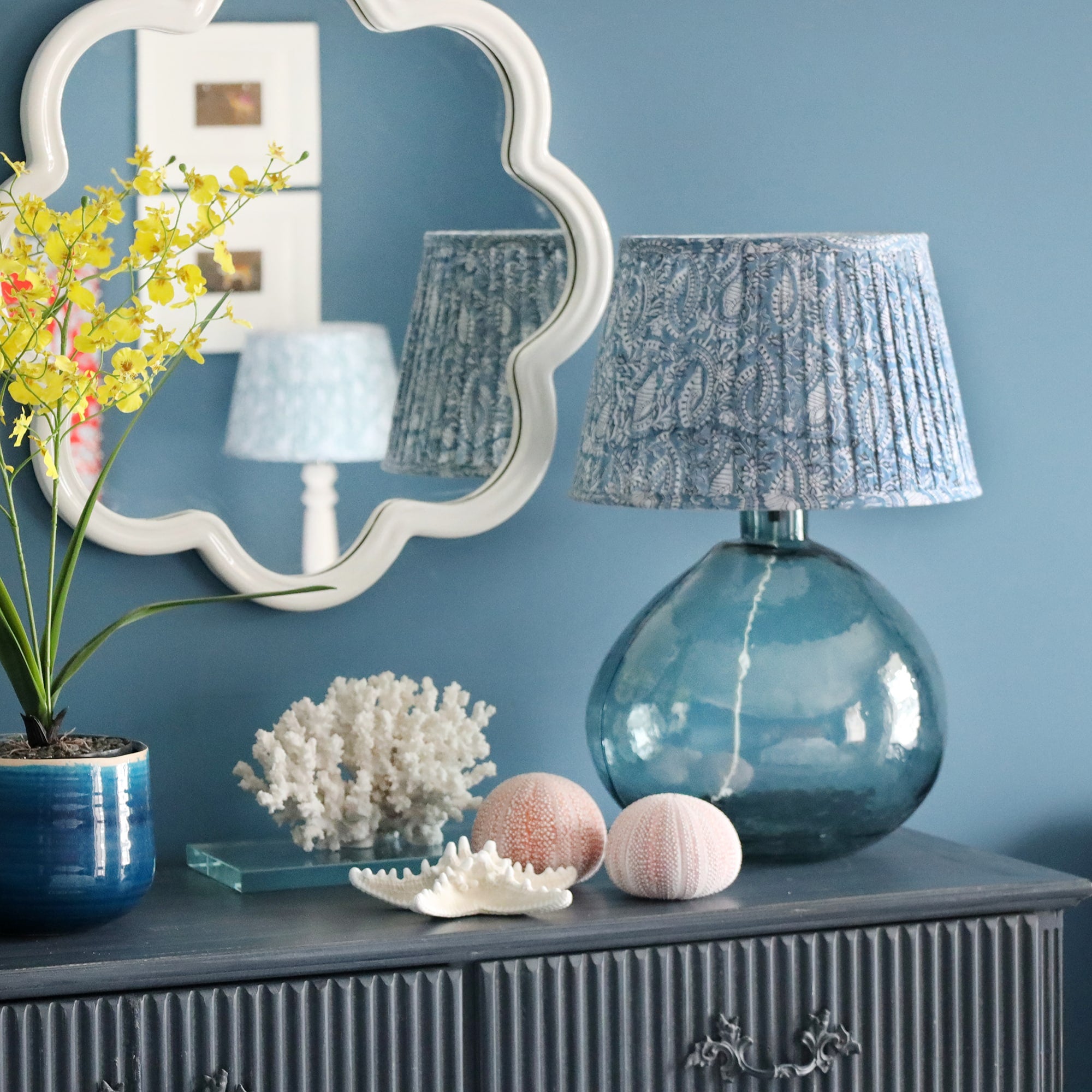 Large Azure paisley pleated lampshade on a seaglass lampbase placed on a sideboard.On the sideboard are various seashells and a plant in a blue pot,in the background is a white wave mirror on the wall