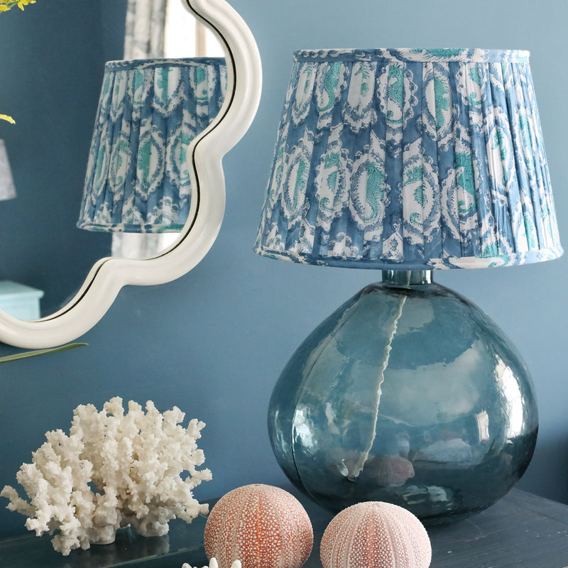 Marine Seahorse pleated lampshade on our Seaglass lampbase on a sideboard.Also on the sideboard are shells and on the wall behind is a white wave mirror